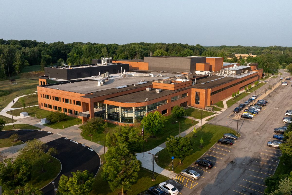 Aerial view of a large, multi-story red brick building with gray roofing, surrounded by green lawns, pathways, and a parking lot with cars.